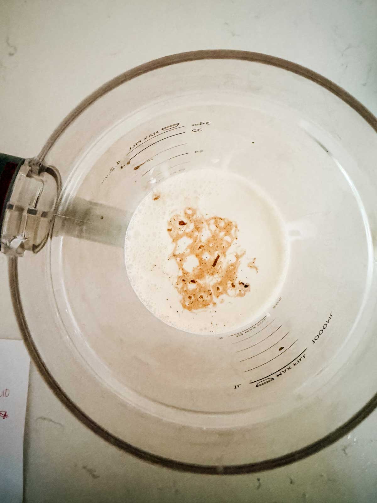 A glass measuring cup filled with ice cream base on a white surface, with measurement markings visible and a metal spoon partially inside the cup.