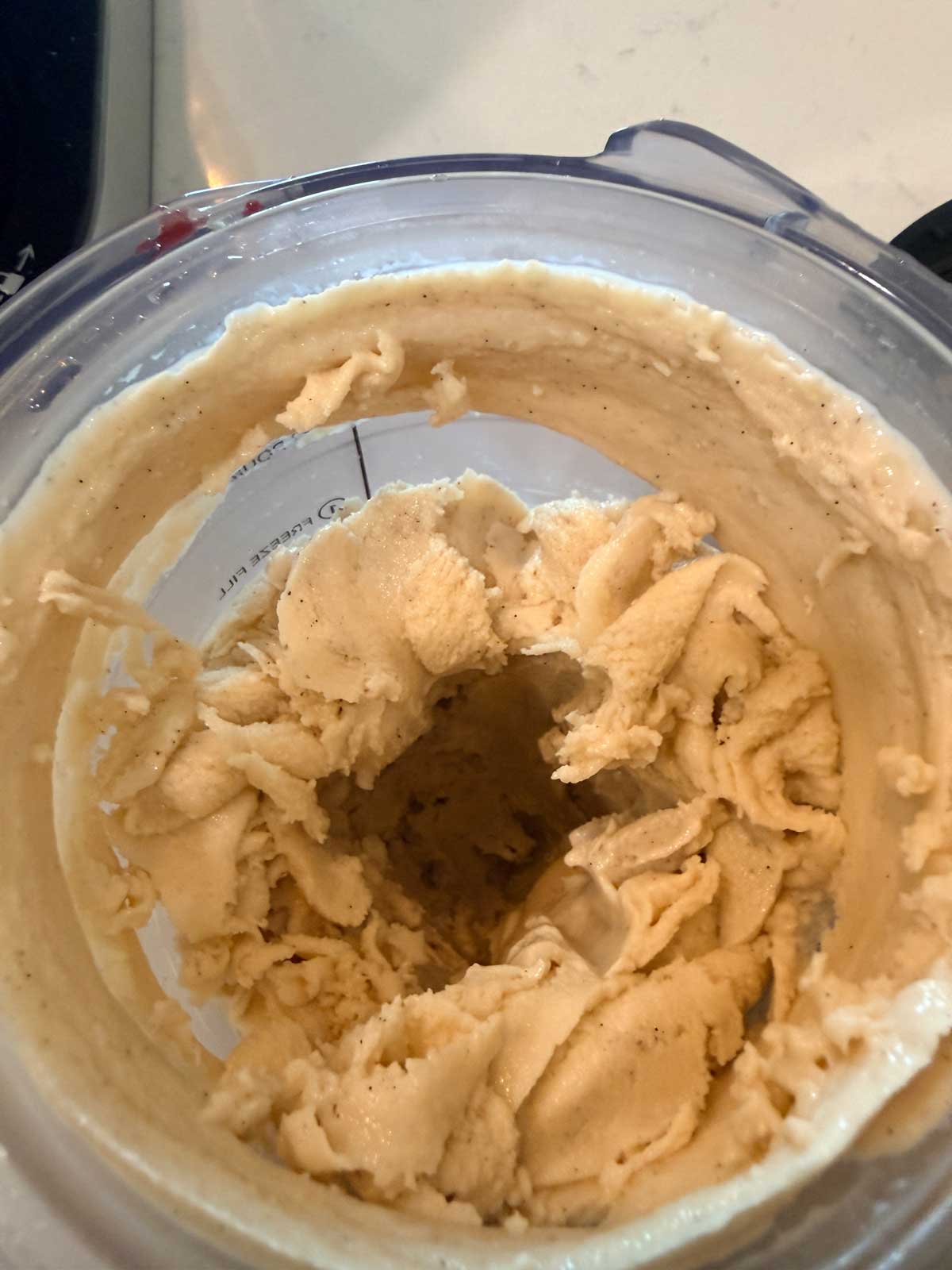 A close-up view of creamy, light beige ice cream or frozen dessert mixture in a blender, with swirled texture and a hole in the center.