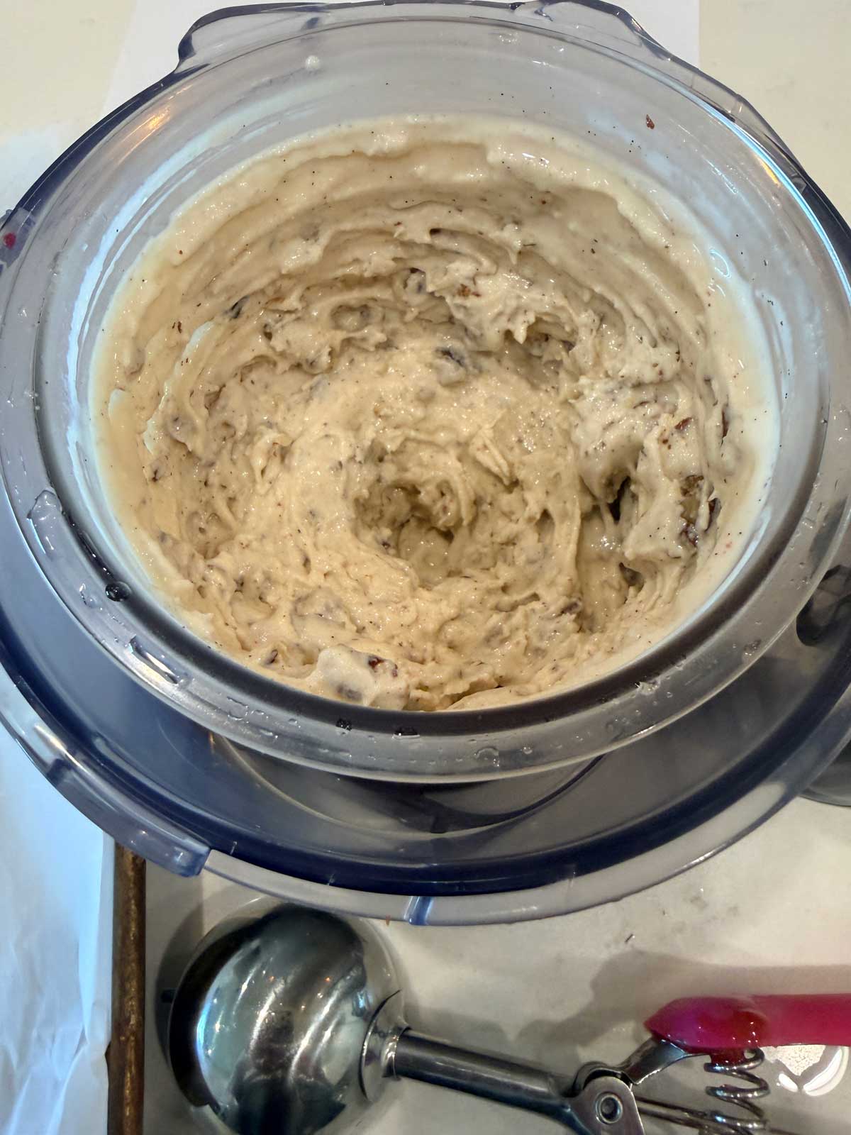 A close-up of freshly churned ice cream in an ice cream maker, with creamy, swirled texture. An ice cream scoop and tongs are placed nearby on the white surface.