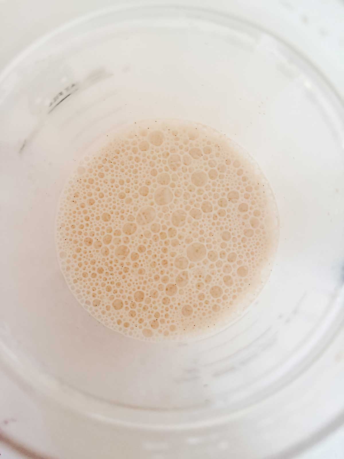 A close-up top view of a frothy, light beige liquid in a transparent cup, showing numerous small bubbles on the surface.