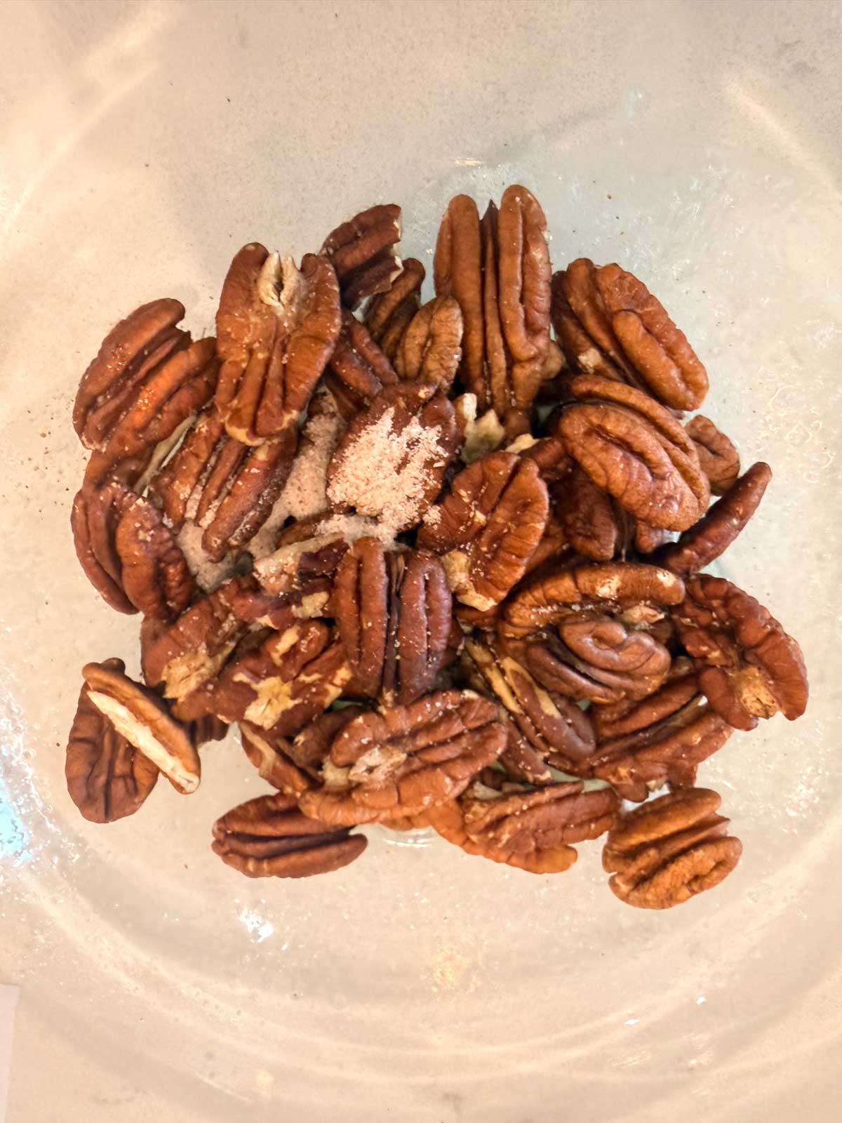 A pile of pecan halves sprinkled with salt and spices sits in a clear bowl on a light-colored surface.