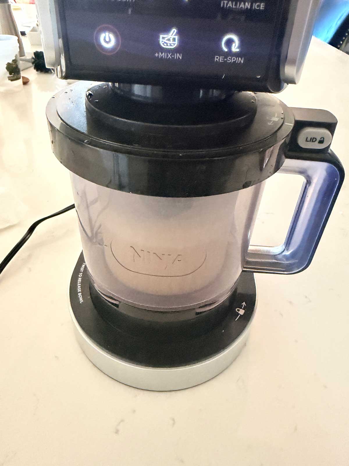 A Ninja ice cream maker sits on a kitchen counter, with its clear container attached and control panel display showing options like Italian Ice, Mix-In, and Re-Spin.