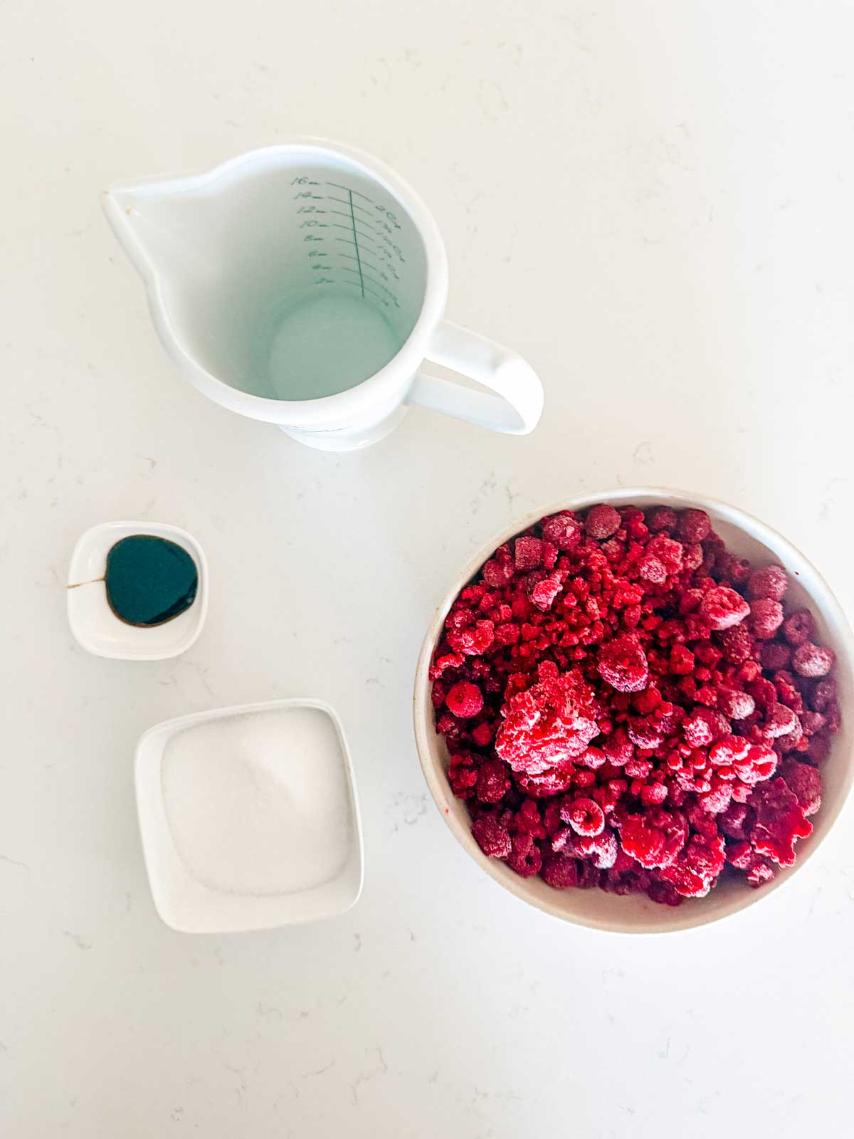 A bowl of frozen raspberries, a measuring cup with water, a small dish of sugar, and a small dish with a green liquid, all arranged on a white surface.