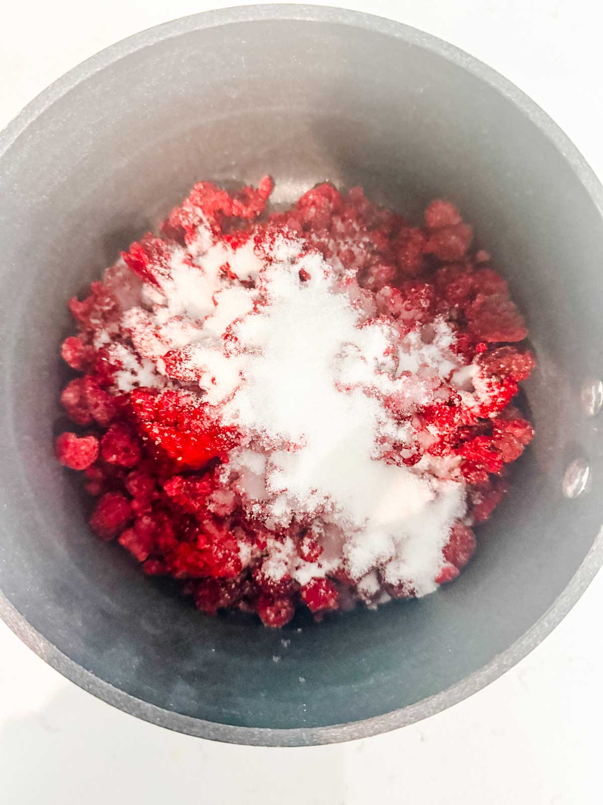 A close-up of a black saucepan containing red berries topped with a pile of white granulated sugar, ready to be cooked.