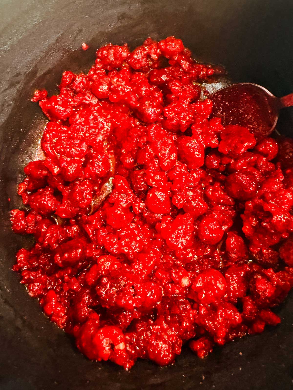 Bright red berries are clustered together in a dark pan, with a spoon partially visible on the right side, coated in the glossy red mixture.
