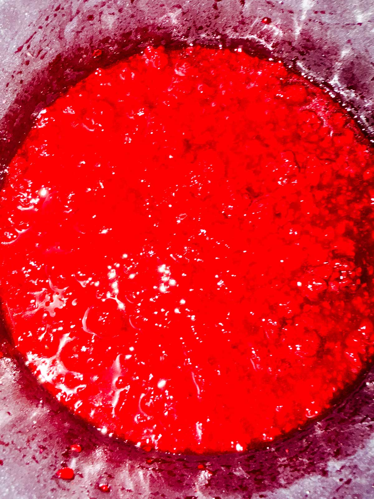 Bright red raspberries that have been cooked down in a saucepan. The edges of the container are visible and stained.