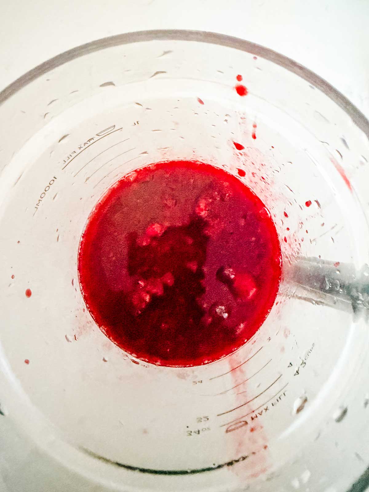 Overhead view of a blender containing a bright red, slightly chunky liquid mixture, with a metal spoon partially visible to the right inside the cup.