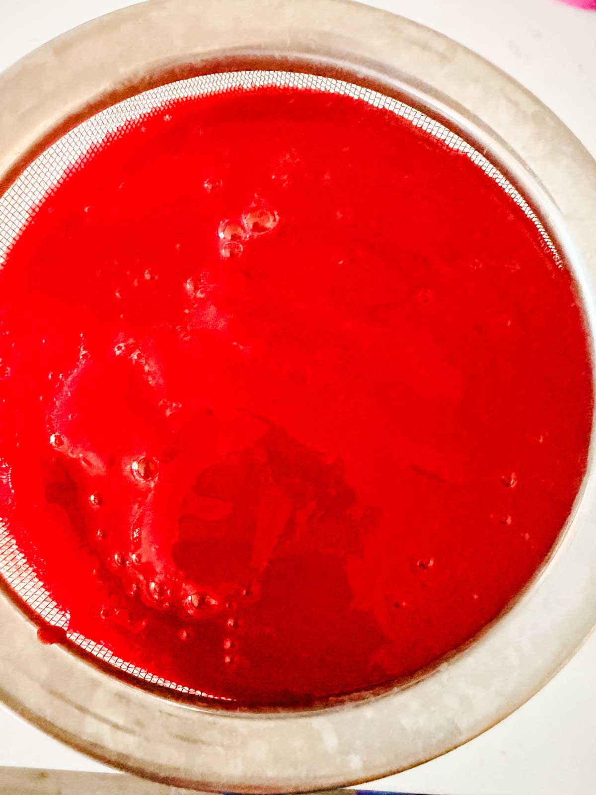A close-up of a metal strainer holding bright raspberry puree.