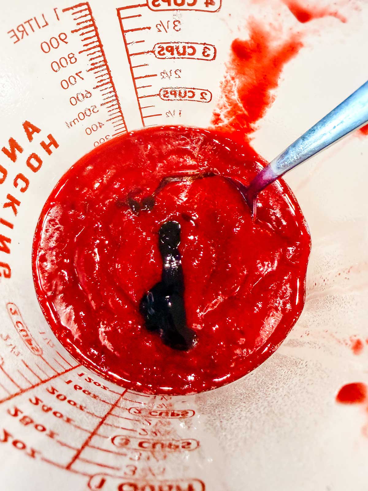 A close-up of a glass measuring cup containing thick red sauce or puree with a streak of dark liquid on top and a metal spoon resting inside. Measurement markings are visible on the cup.