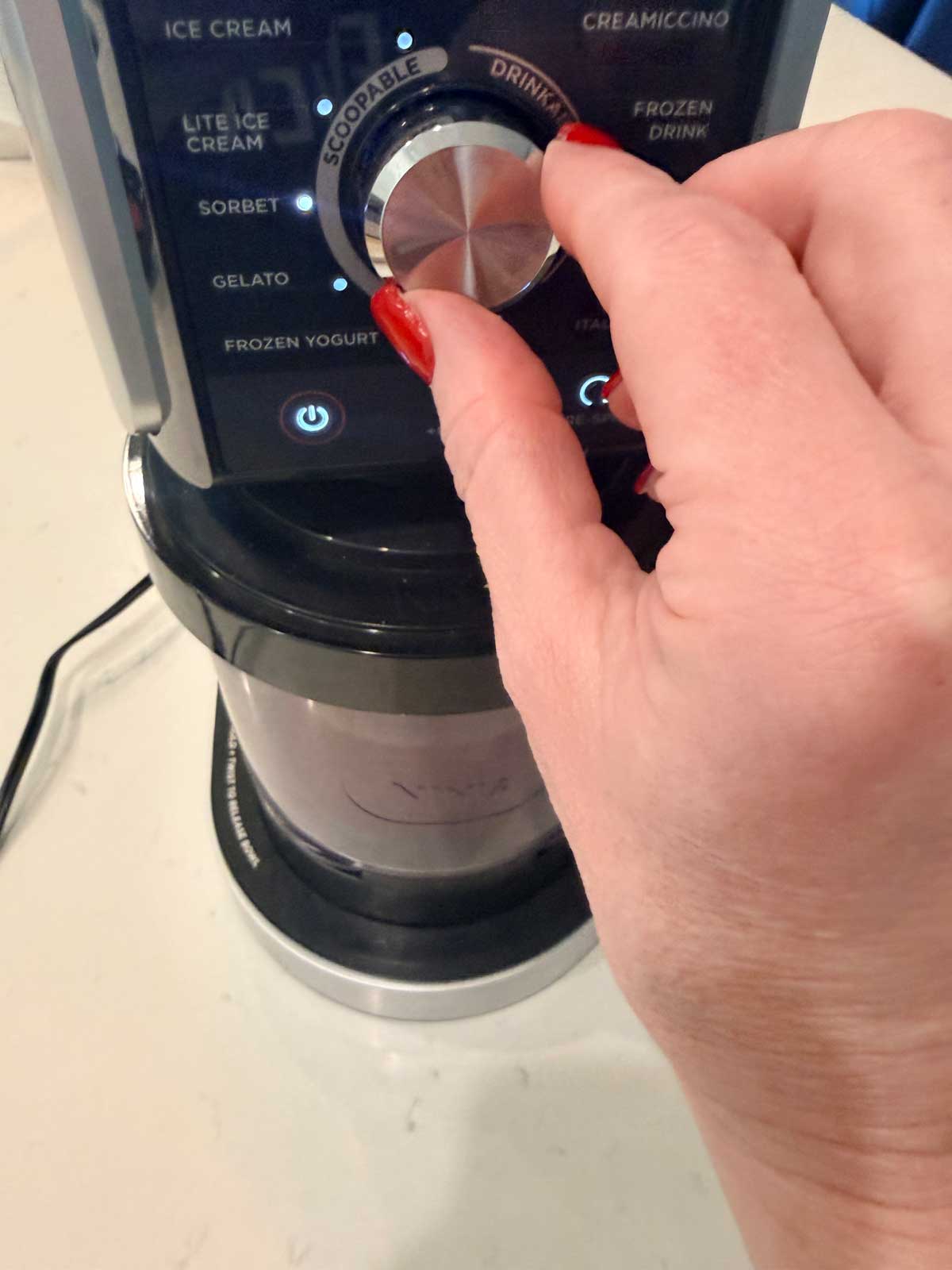 A hand with red nail polish turns the dial on an ice cream maker, selecting the Drink mode from various options like ice cream, gelato, and frozen yogurt.
