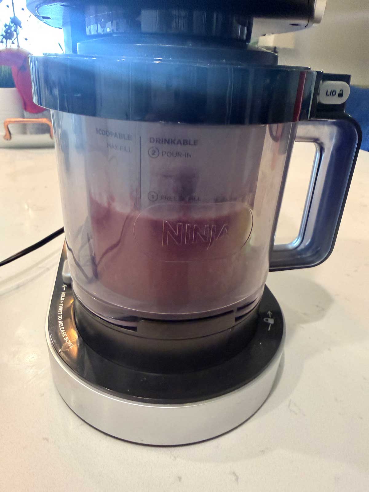 A close-up of a Ninja blender containing a pink mixture, likely being blended, sits on a kitchen counter with some appliances visible in the background.