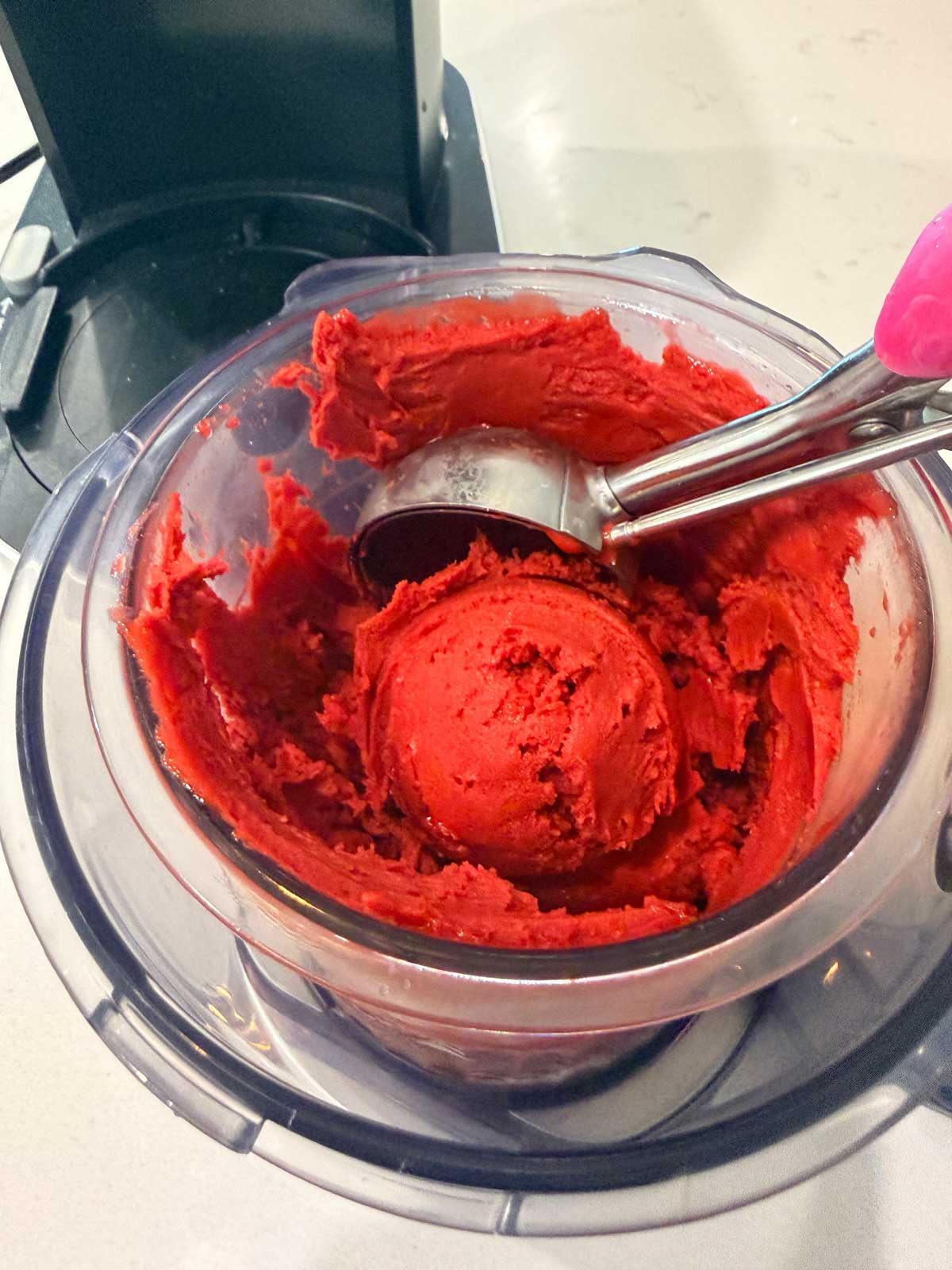 A metal scoop is lifting a ball of bright red sorbet from a clear food processor bowl, which is sitting on a white countertop.