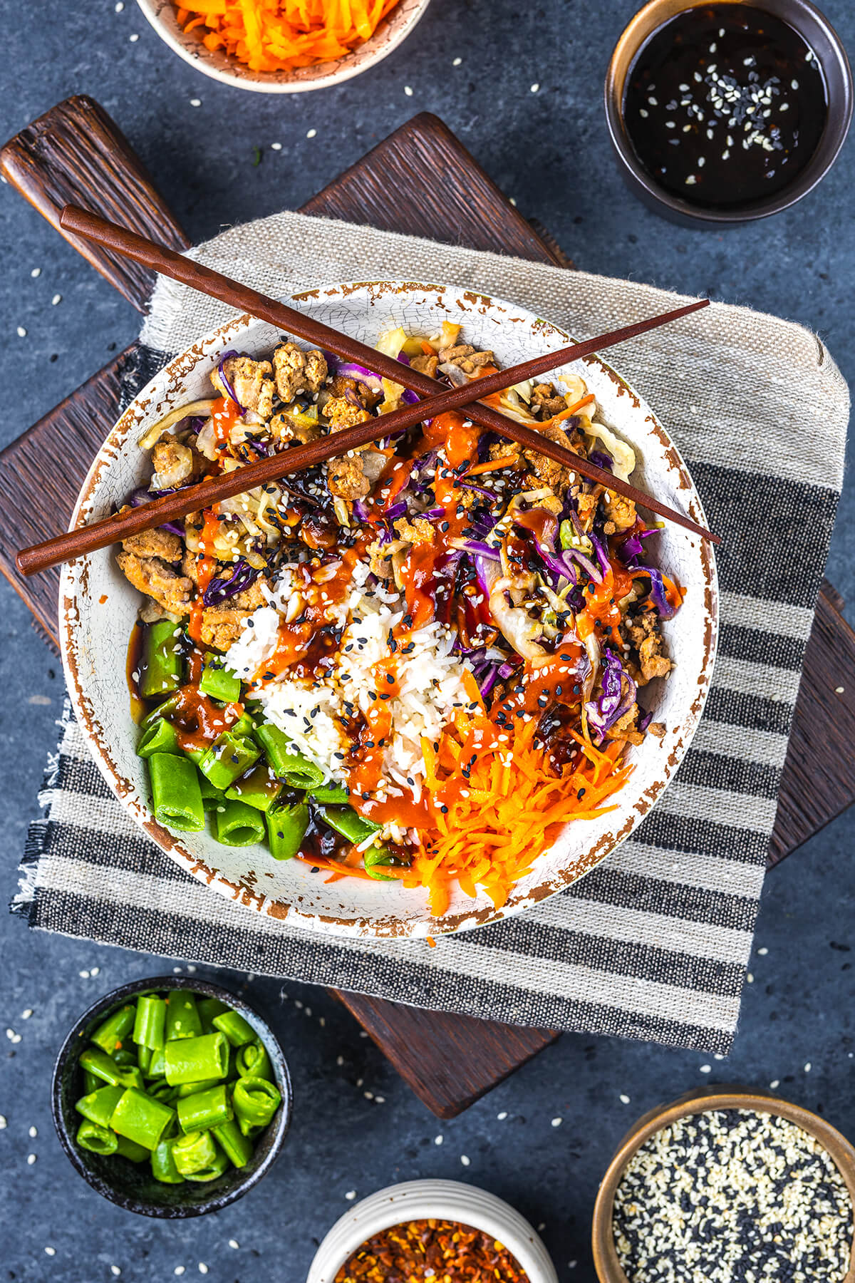 A bowl filled with rice, shredded carrots, snap peas, purple cabbage, and grilled chicken, topped with sauce and sesame seeds, with chopsticks resting on top. Surrounding bowls contain sauce, sesame seeds, snap peas, and chili flakes.