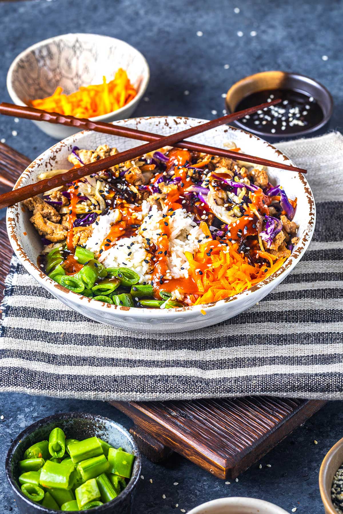 A bowl of rice topped with shredded carrots, green beans, purple cabbage, and a saucy protein, garnished with black sesame seeds. Chopsticks rest on top, with small bowls of sauce and vegetables nearby.