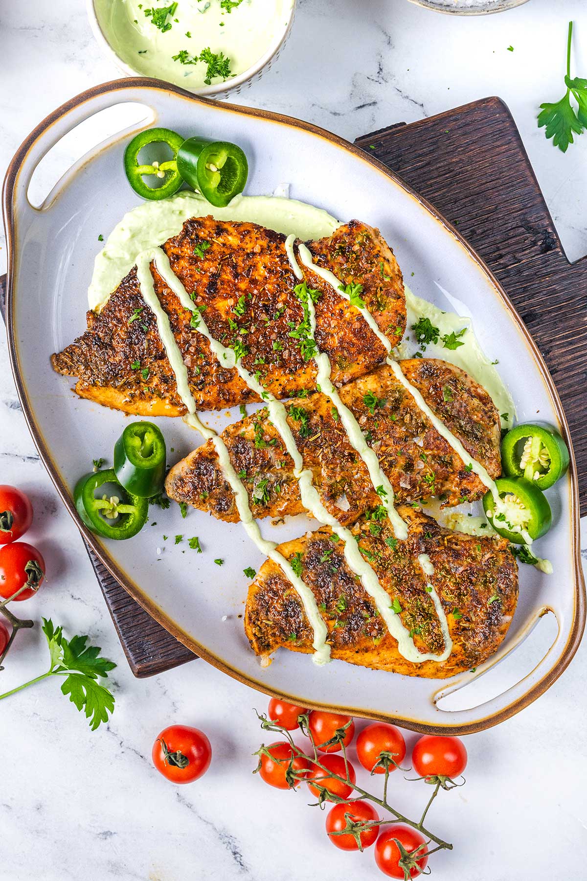 A serving platter with sliced, seasoned blackened chicken drizzled with a creamy sauce, garnished with fresh parsley and sliced jalape&ntilde;os, surrounded by cherry tomatoes and parsley on a white marble surface.