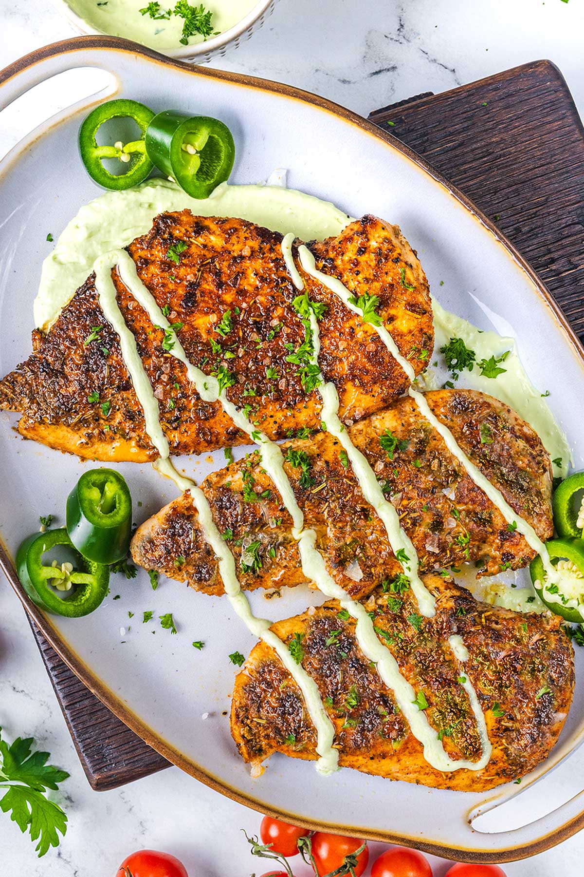 A platter of blackened chicken breasts garnished with chopped herbs, drizzled with a creamy white sauce, served atop a vibrant green sauce, accompanied by sliced jalapeños and fresh parsley on the side.
