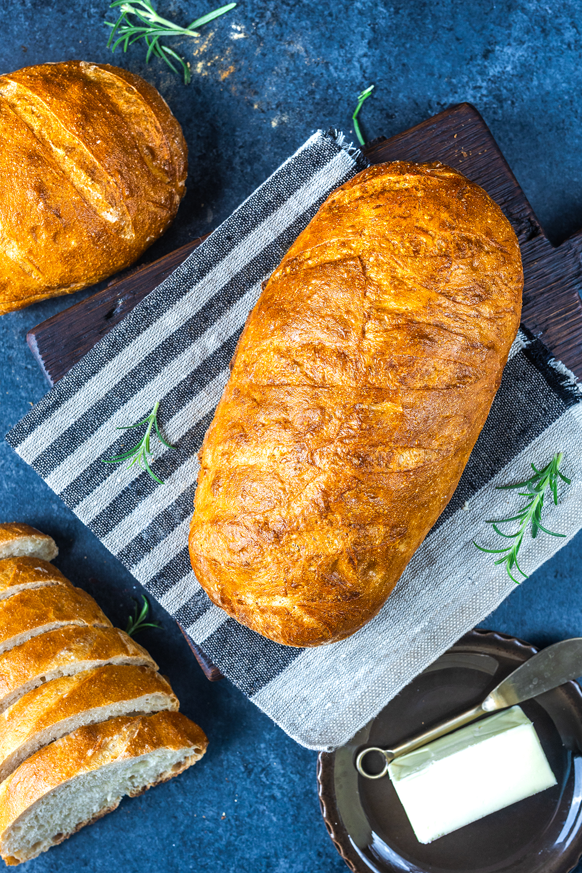 A golden, freshly baked loaf of bread rests on a striped towel with sliced bread, a butter dish, and sprigs of rosemary nearby, all arranged on a dark blue surface.