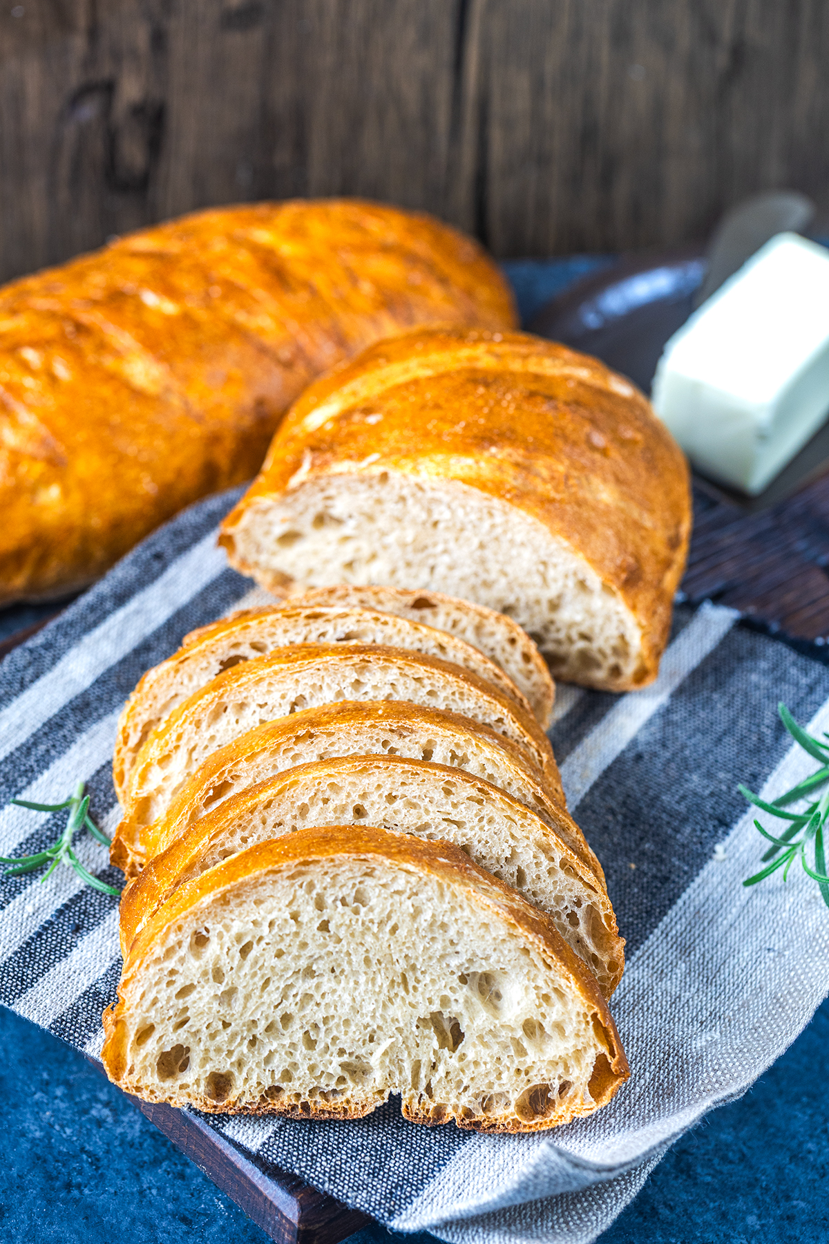 A loaf of rustic bread, partially sliced, is displayed on a striped cloth with a stick of butter and sprigs of rosemary nearby, set on a dark wooden surface.