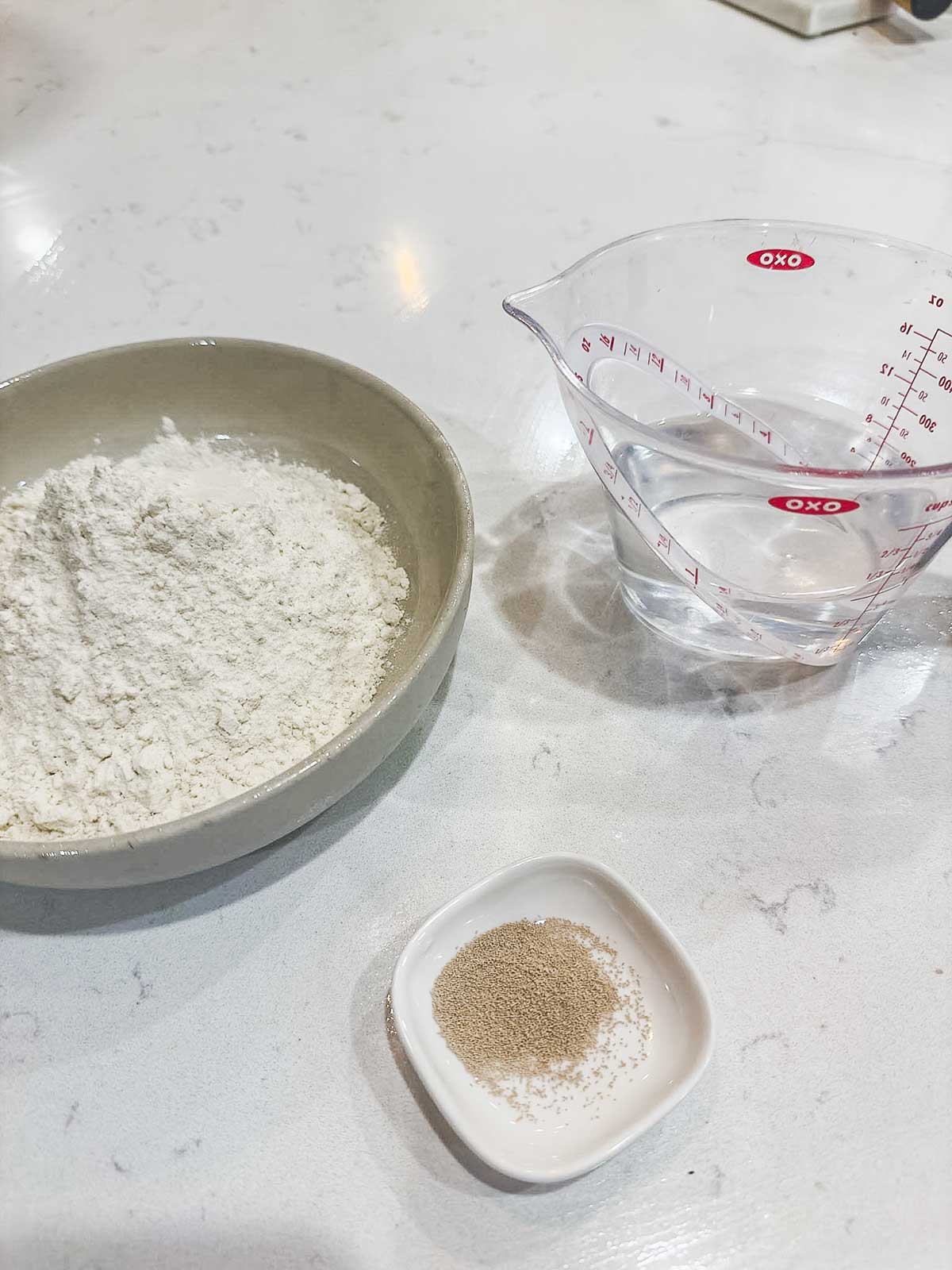 A bowl of flour, a small dish of dry yeast, and a measuring cup of water are arranged on a white countertop.