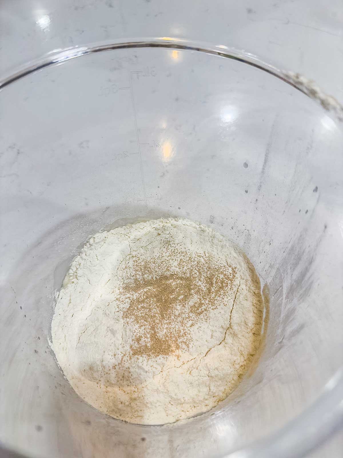 A clear mixing bowl containing a small mound of flour with a sprinkling of yeast on top, placed on a light-colored surface.