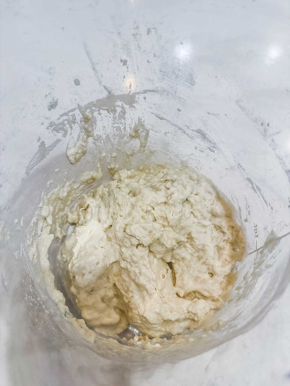 A close-up of sticky bread dough in a clear mixing bowl with remnants of flour and dough on the sides, placed on a white countertop.