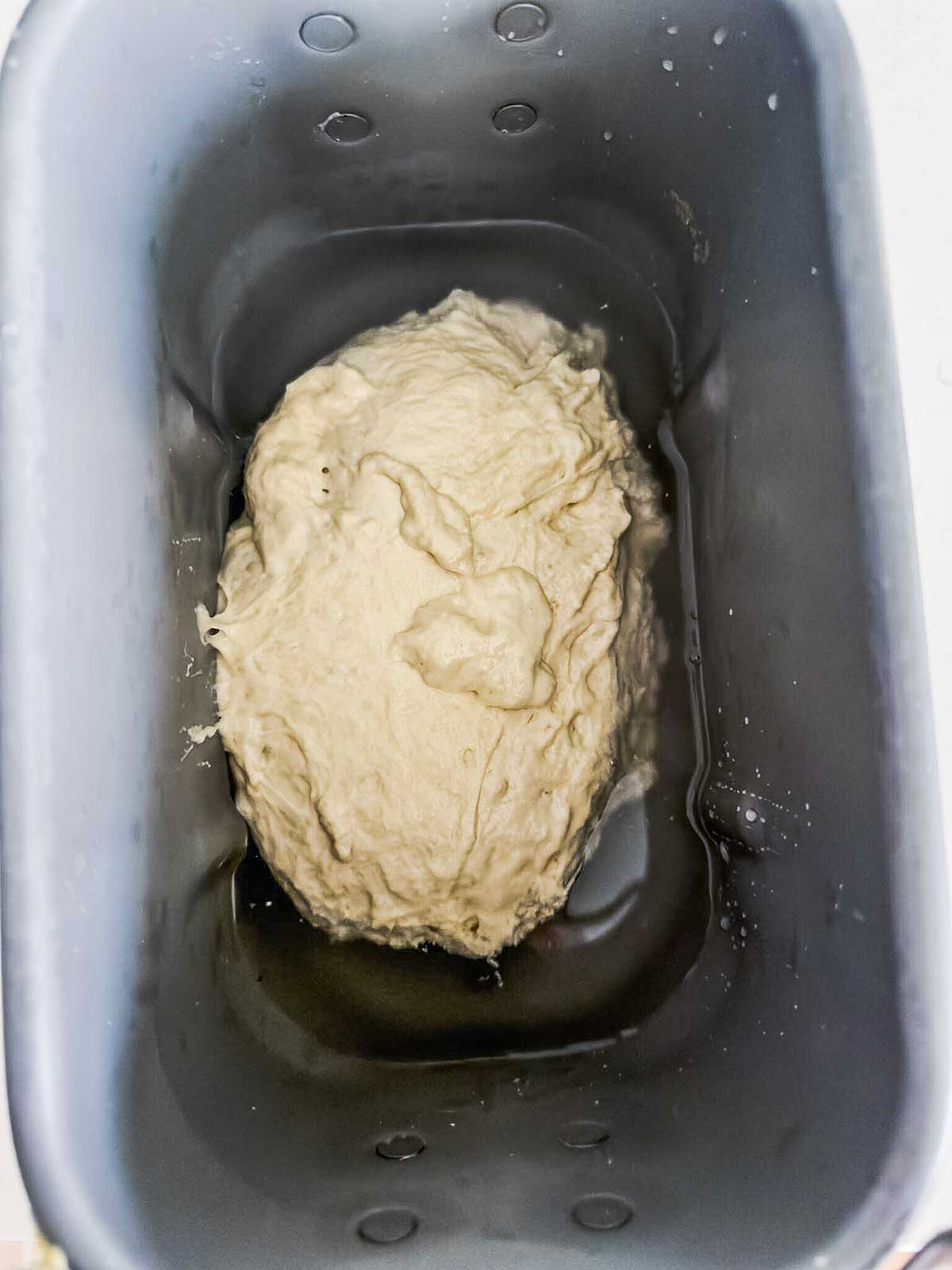 A ball of dough sits inside a bread machine pan, ready for kneading or rising. The pan is metallic and has droplets of water on its sides.