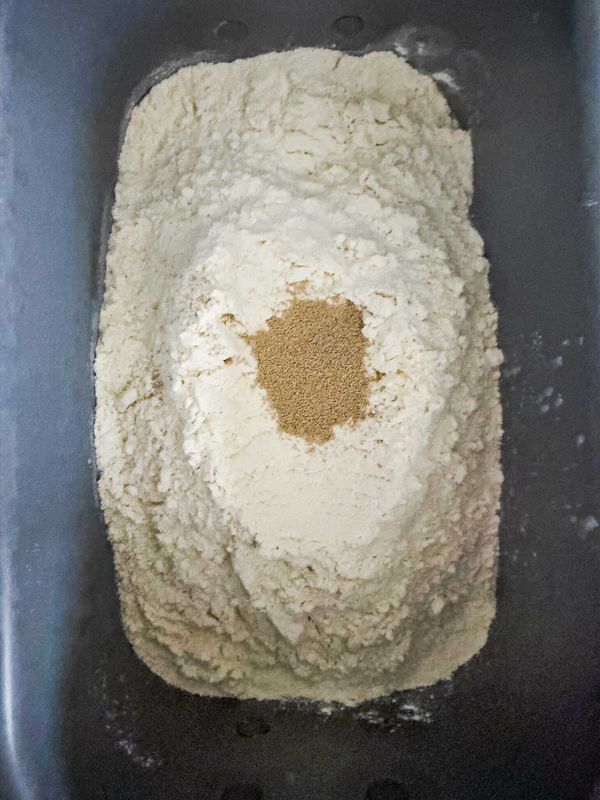 A mound of flour with a well in the center filled with dry yeast, placed inside a rectangular container, ready for baking preparation.