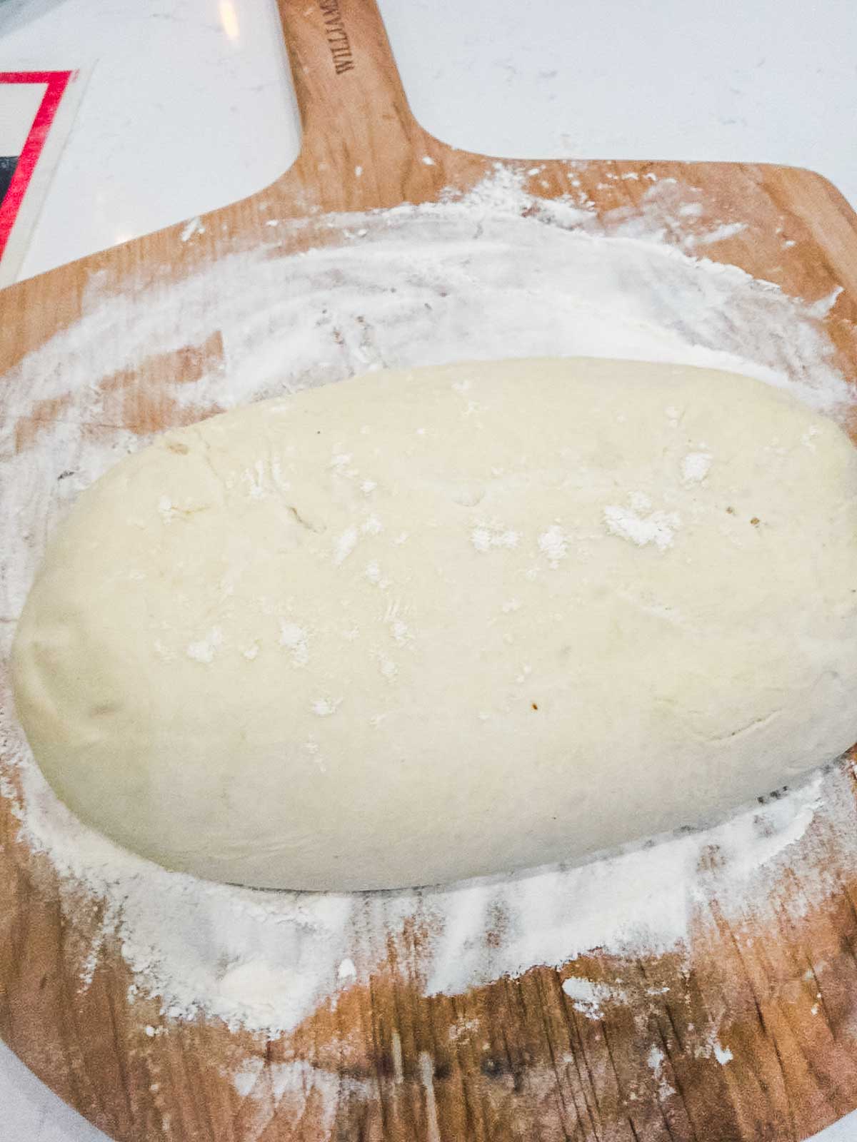 A loaf-shaped ball of dough rests on a wooden pizza peel dusted with flour, ready for baking.