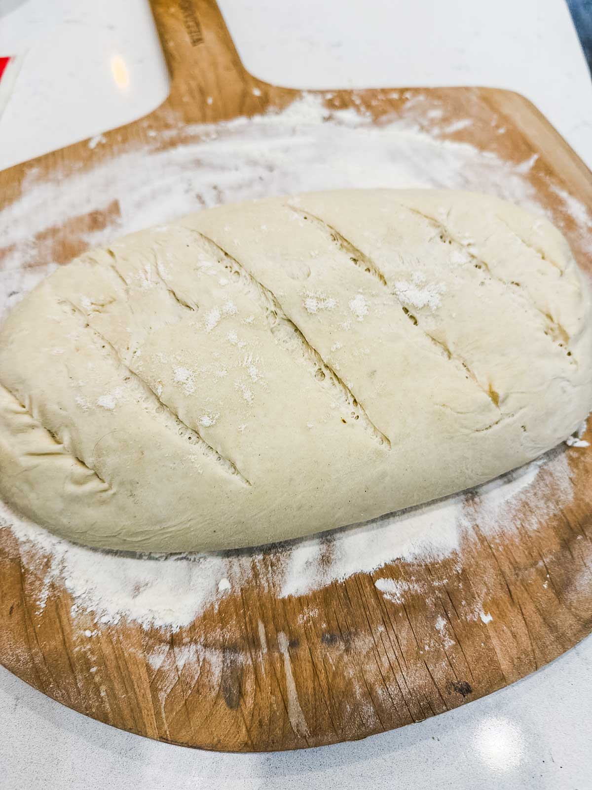 An unbaked loaf of bread dough with slits on top sits on a floured wooden pizza peel, ready for baking.