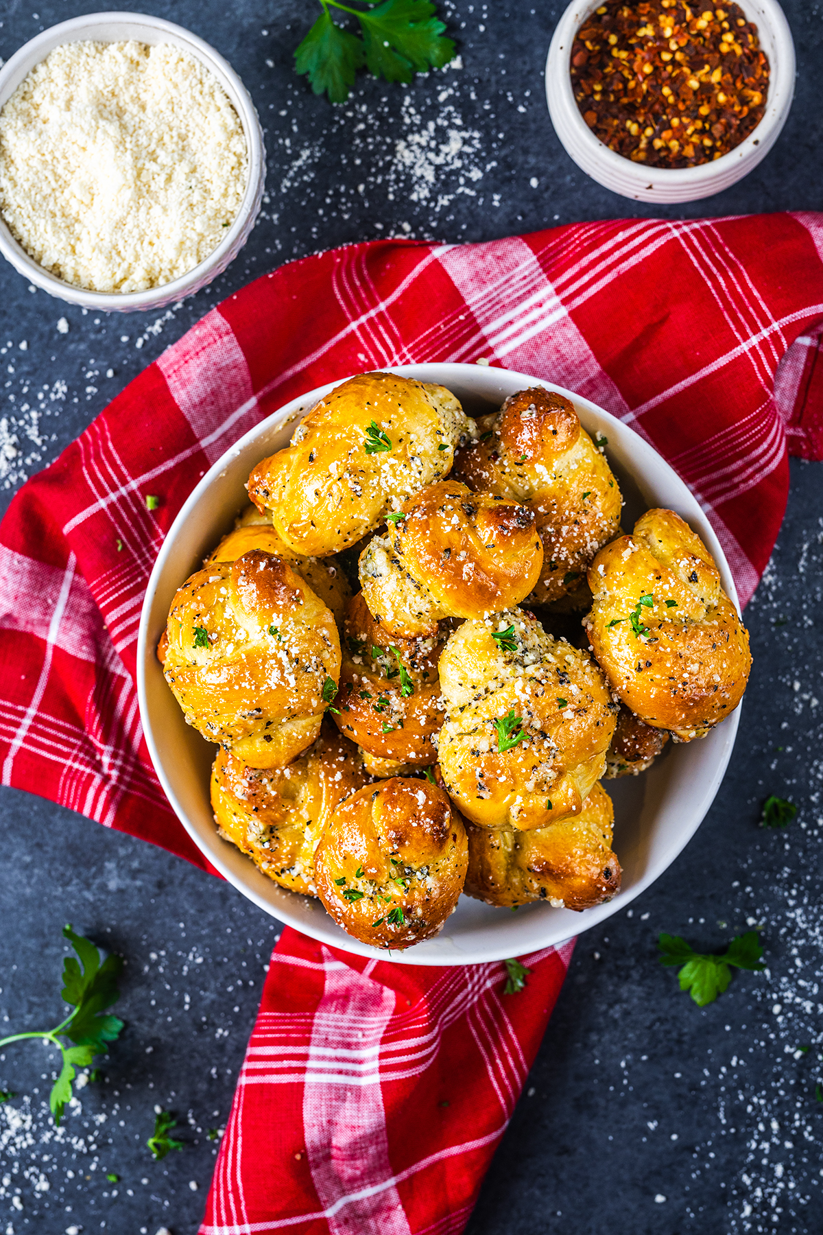 A white bowl filled with golden garlic knots topped with herbs and cheese sits on a red plaid cloth. Parmesan cheese and red pepper flakes are in small bowls nearby. Parsley garnishes are scattered around.