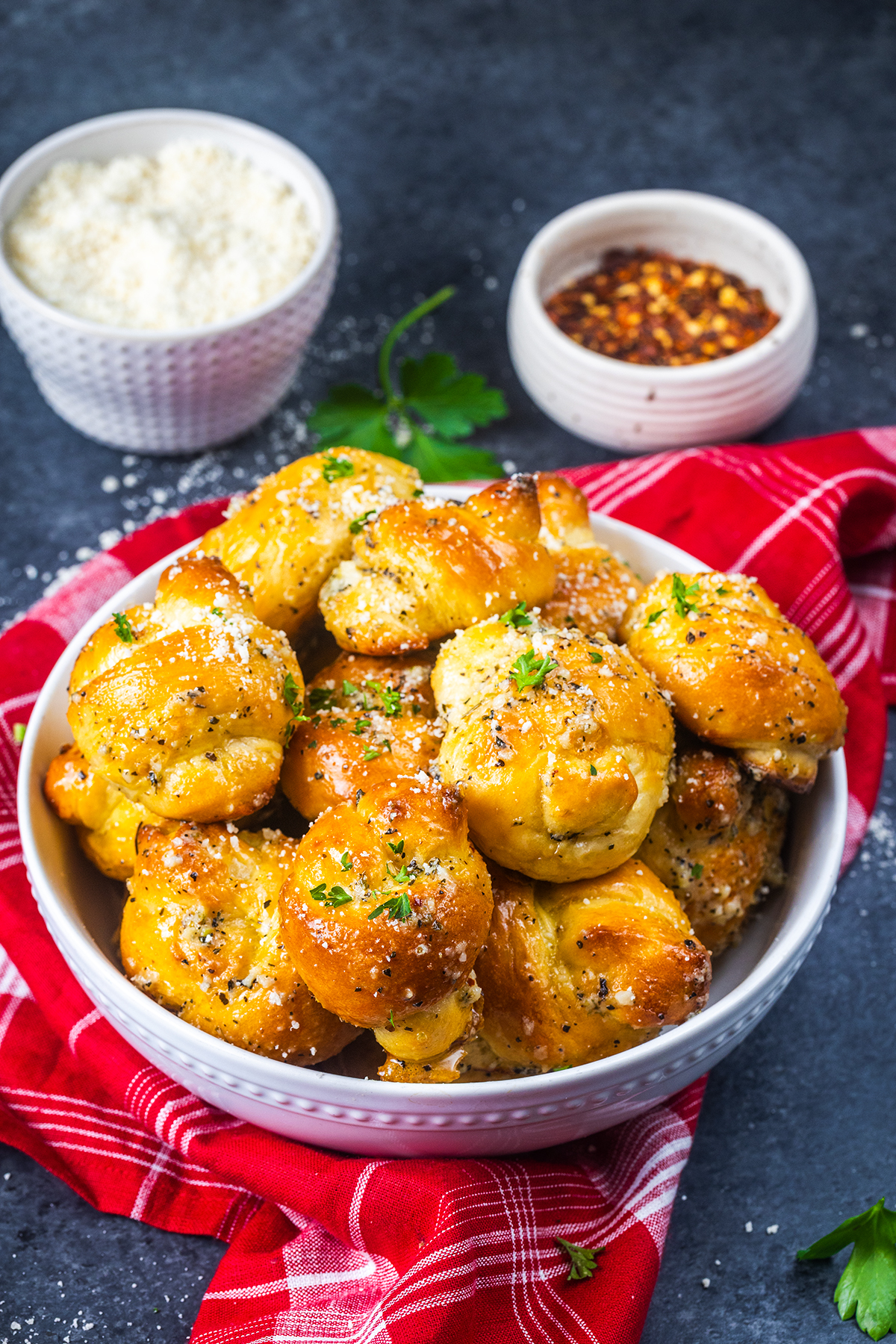 A white bowl filled with garlic knots topped with herbs and cheese, placed on a red cloth. In the background, small bowls contain grated cheese and red pepper flakes.