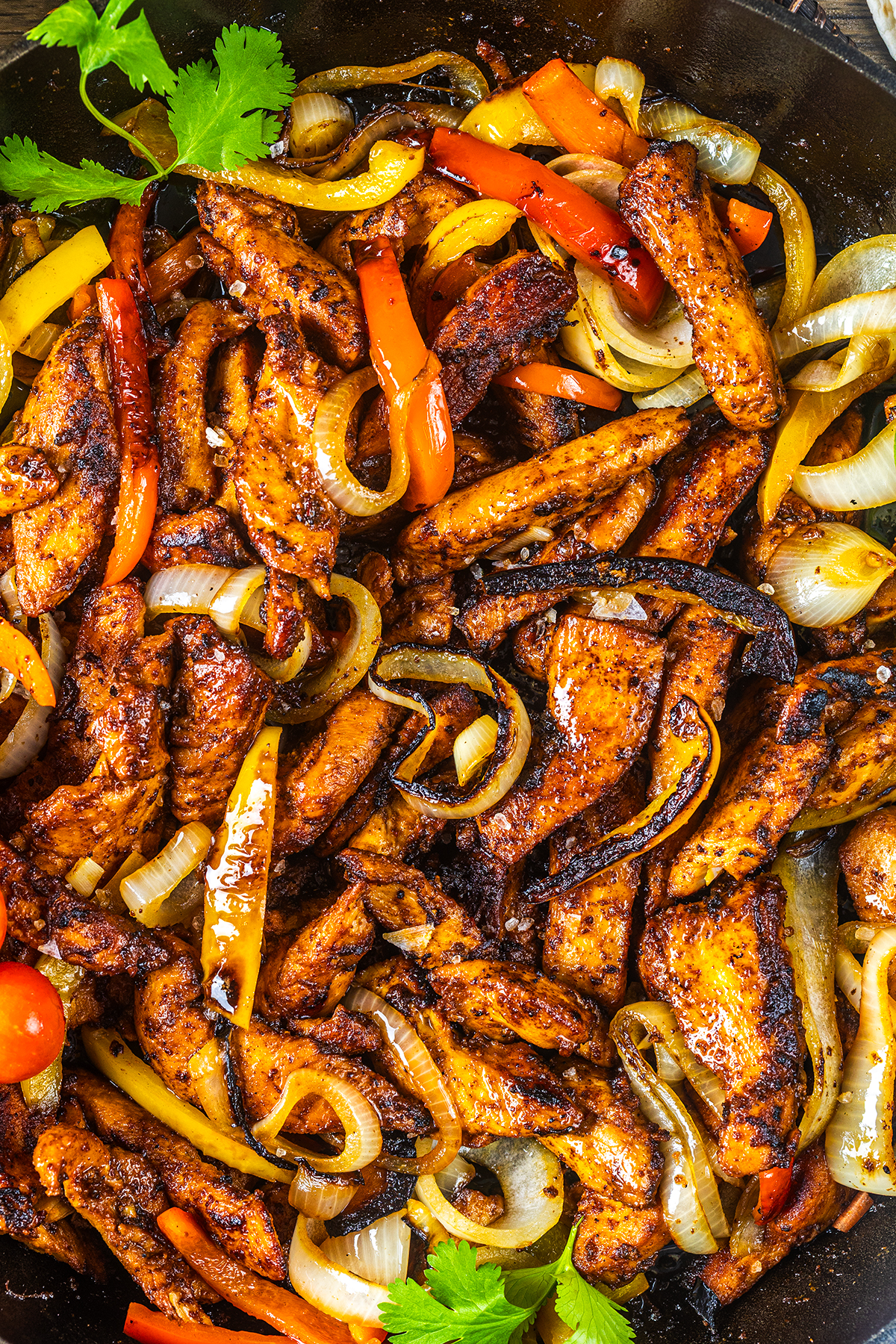 Colorful chicken fajitas with grilled strips of spiced chicken, sautéed bell peppers in yellow and red, and onions, garnished with fresh cilantro in a skillet. The dish appears hot and flavorful.