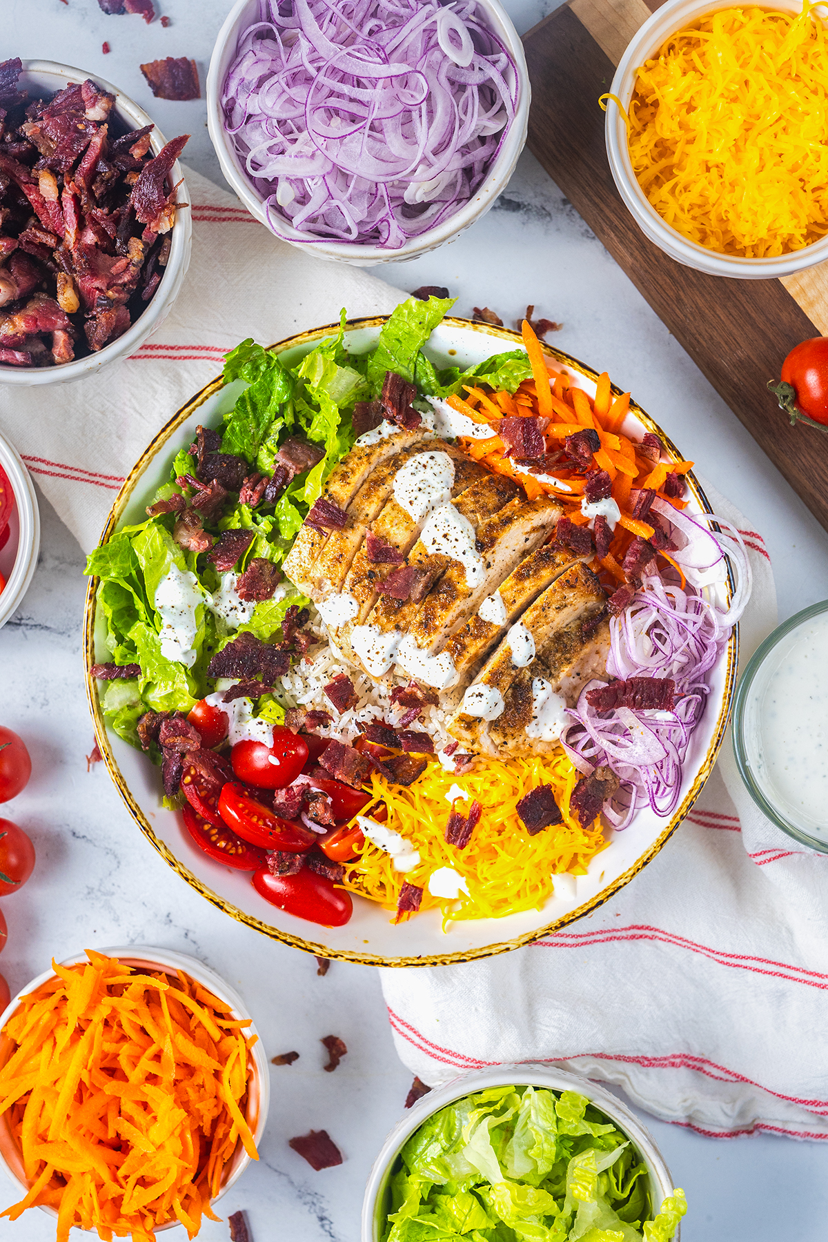 A colorful salad bowl with sliced grilled chicken, shredded cheese, bacon bits, cherry tomatoes, shredded carrots, red onions, lettuce, and ranch dressing. Surrounding bowls hold extra toppings on a white table.