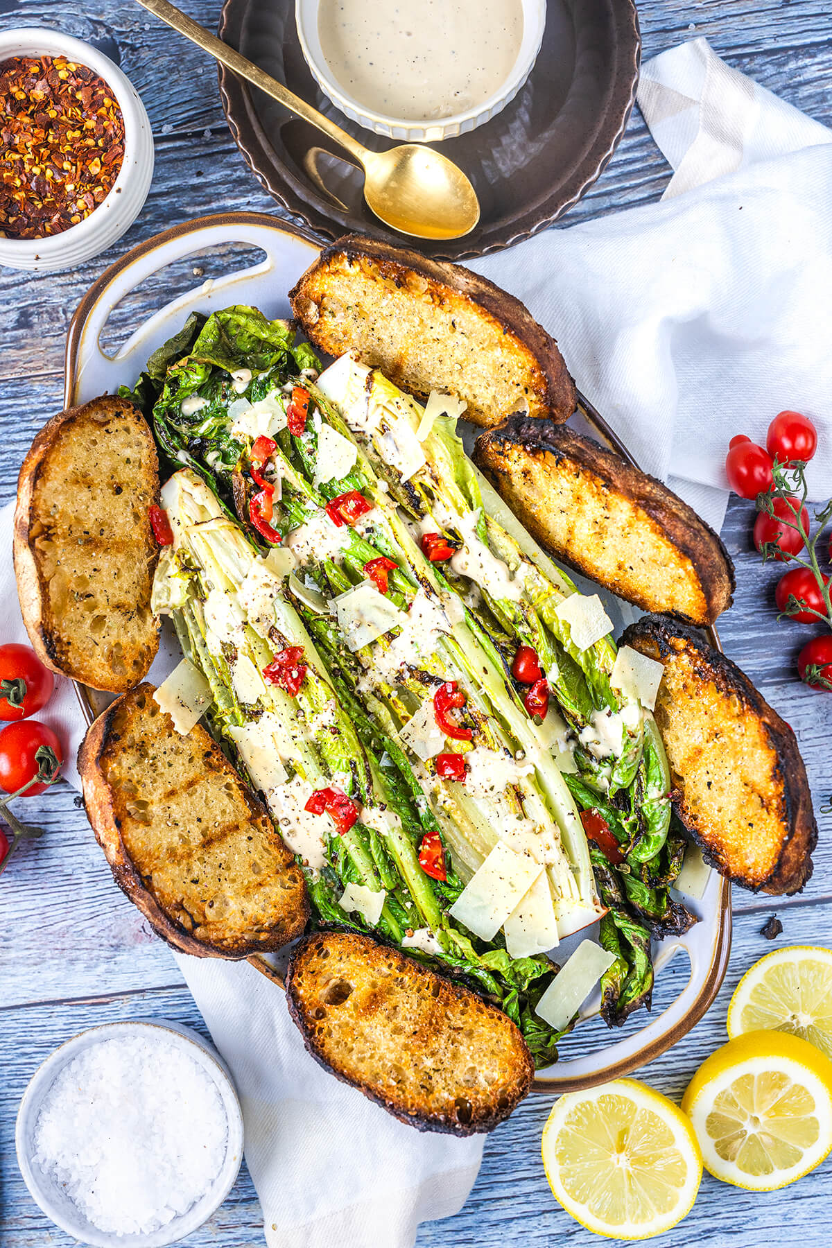 A platter of grilled romaine lettuce topped with shaved cheese and red chili pieces, surrounded by toasted bread slices. Nearby are lemon slices, cherry tomatoes, a bowl of dressing, red pepper flakes, and a bowl of salt.