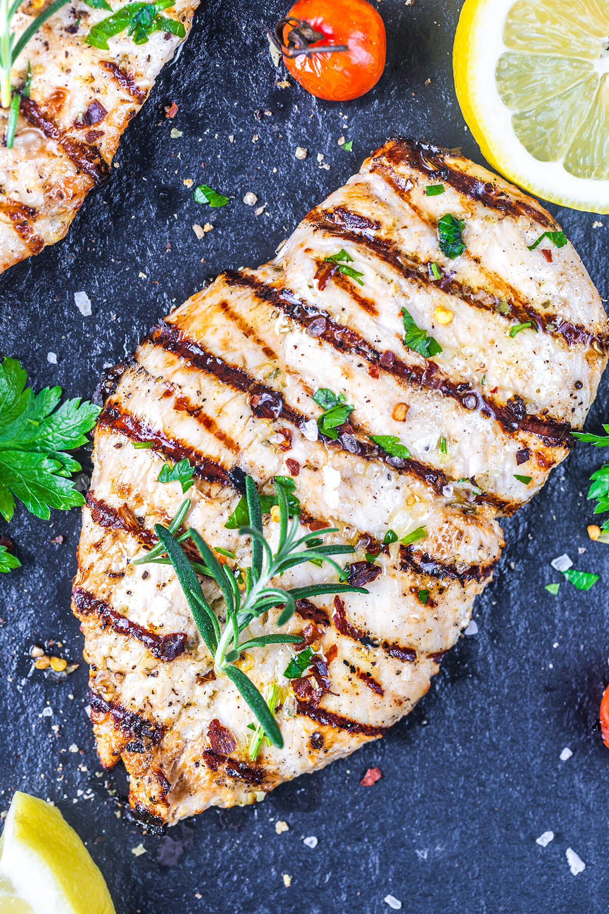 Grilled chicken breast garnished with fresh herbs and coarse salt, served with cherry tomatoes, parsley, and lemon wedges on a dark slate surface.