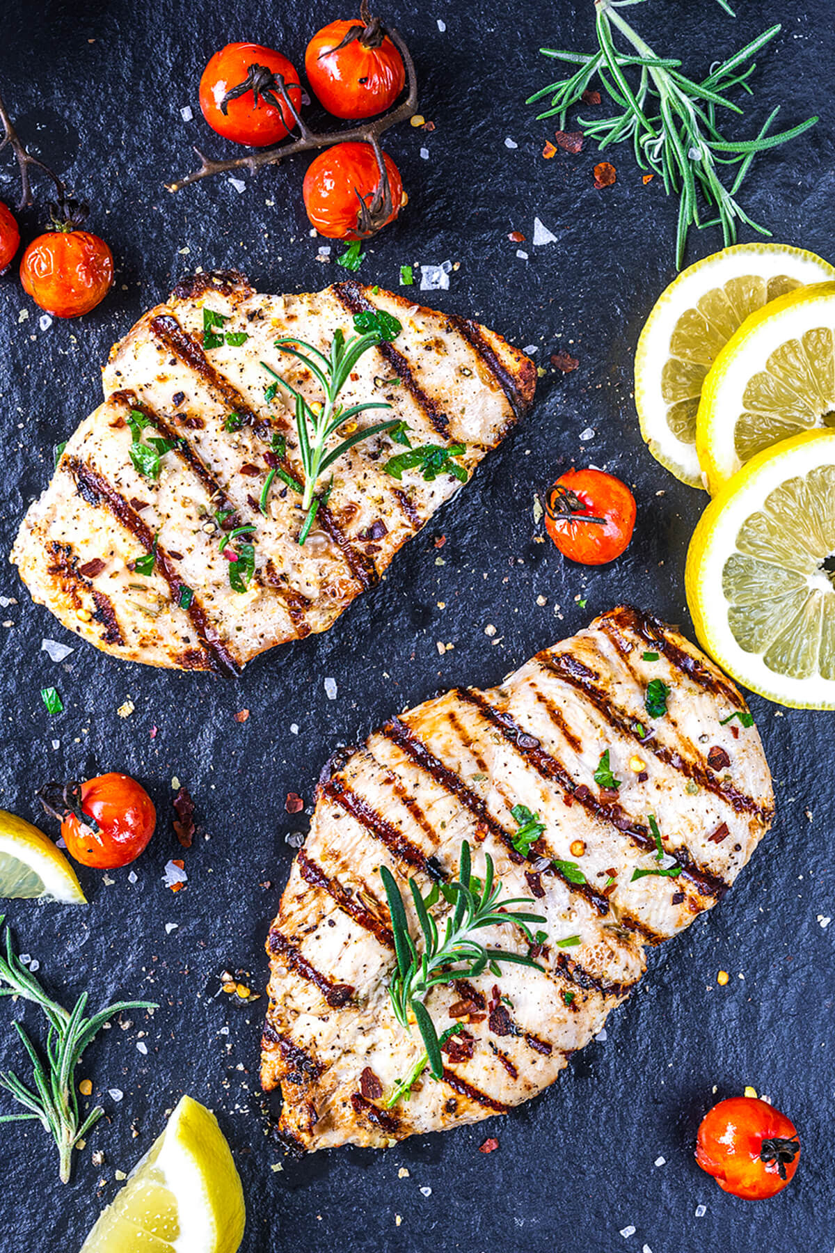Two grilled chicken breasts with grill marks are garnished with rosemary and served on a dark surface, surrounded by roasted cherry tomatoes, fresh rosemary sprigs, and lemon slices. Coarse salt and herbs are sprinkled around.