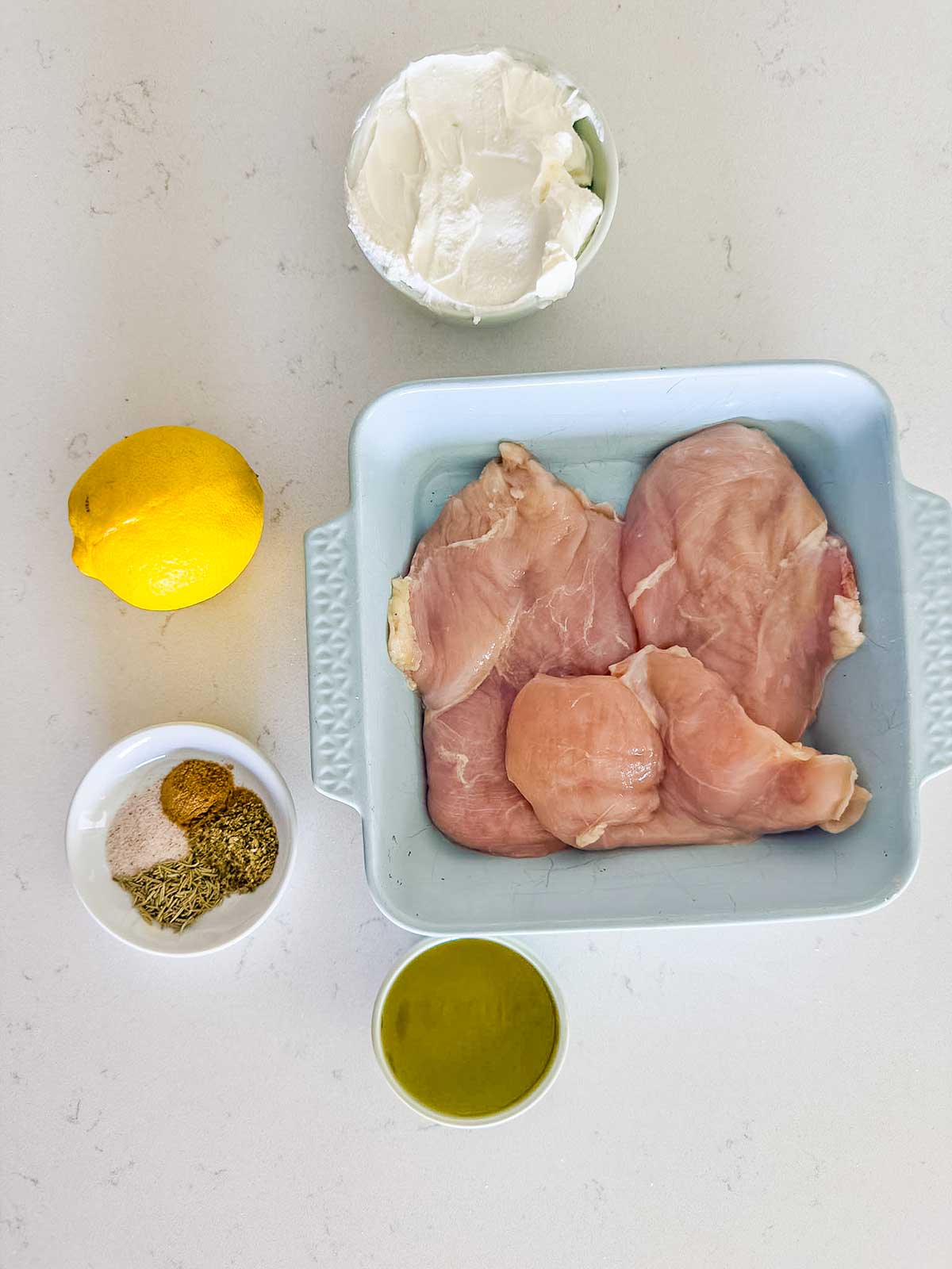 Raw chicken breasts in a light blue baking dish, surrounded by a lemon, a bowl of yogurt, a bowl of spices, and a bowl of olive oil on a white countertop.