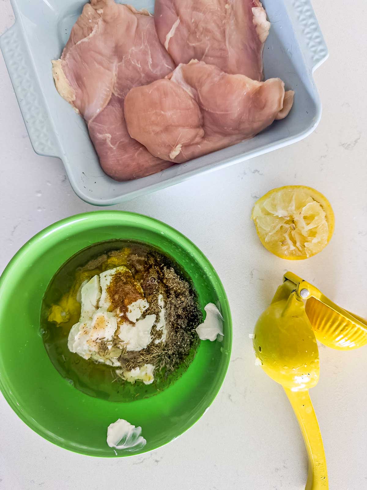 Raw chicken breasts in a baking dish, a squeezed lemon and juicer, and a green bowl with yogurt, spices, herbs, and oil on a white surface, likely for preparing a marinade.