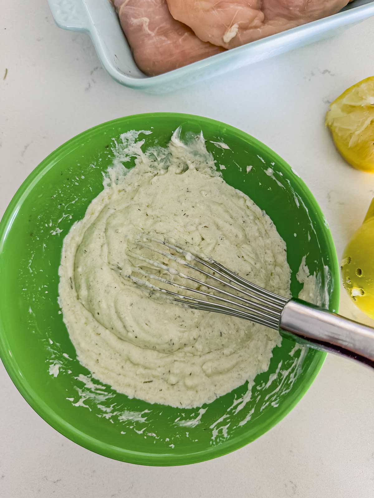 A whisk rests in a green bowl filled with a creamy white mixture; nearby are lemon halves and a dish with raw chicken breasts on a white countertop.