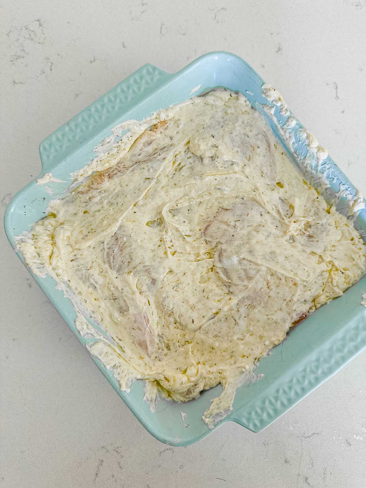 A blue baking dish containing raw chicken coated in a creamy, herbed marinade sits on a light-colored countertop.