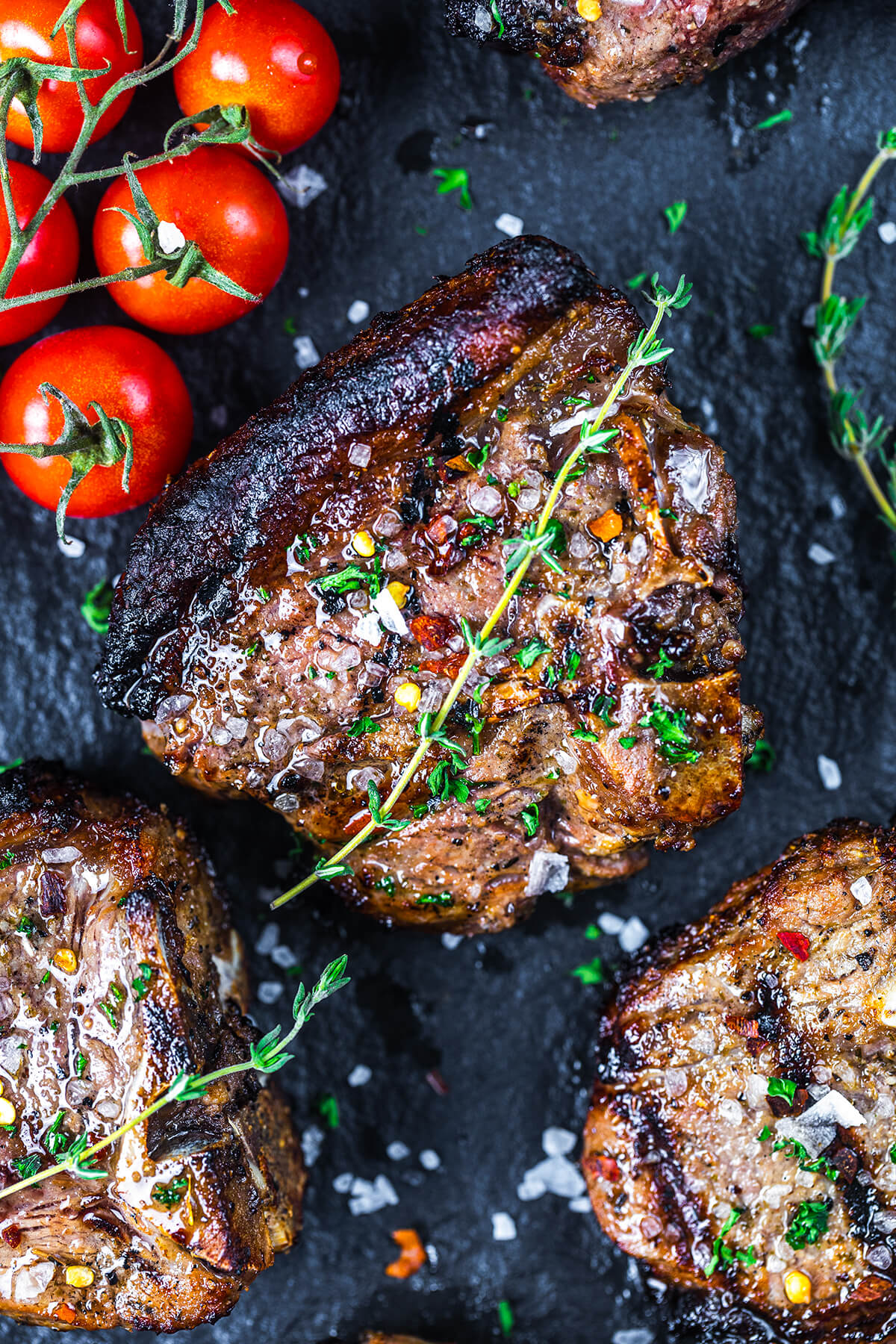 Close-up of grilled lanb pieces garnished with herbs and coarse salt, with a sprig of thyme on top. Cherry tomatoes on the vine are visible in the background, all arranged on a dark slate surface.