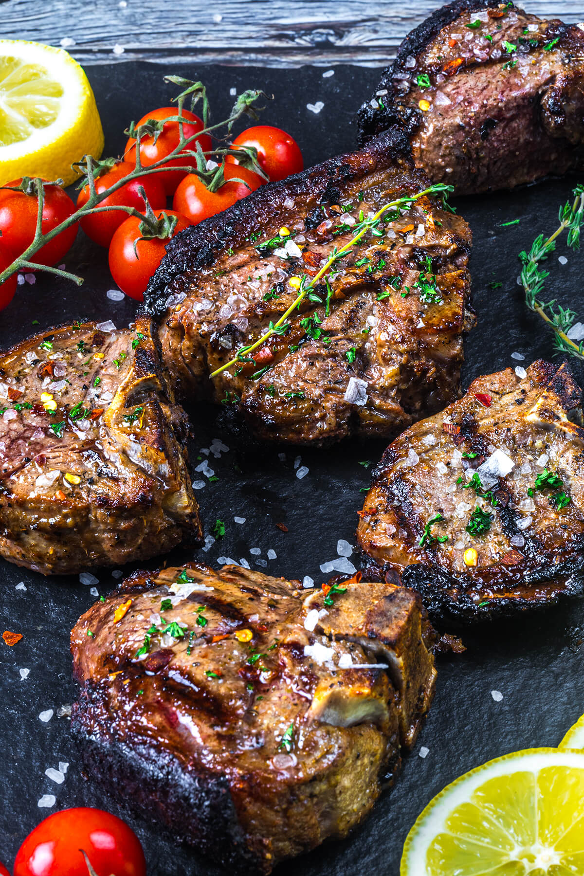 Grilled, seasoned lamb chops garnished with herbs and sea salt on a dark slate, surrounded by cherry tomatoes on the vine and lemon slices.
