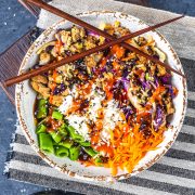 A colorful rice bowl topped with shredded carrots, snap peas, cabbage, crumbled tofu, white rice, sesame seeds, and a drizzle of red sauce, with chopsticks resting across the bowl on a striped placemat.