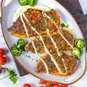 A serving platter with three pieces of blackened chicken breast topped with creamy sauce, garnished with chopped herbs and sliced jalapeños. Cherry tomatoes and parsley surround the platter.