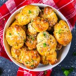 A bowl of golden, baked garlic knots topped with herbs and grated cheese sits on a red plaid cloth, with a few sprigs of parsley scattered around.