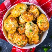 A bowl of golden, baked garlic knots topped with herbs and grated cheese sits on a red plaid cloth, with a few sprigs of parsley scattered around.