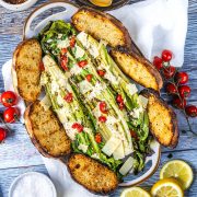 A platter with grilled romaine lettuce topped with parmesan, red peppers, and dressing, surrounded by toasted bread slices. Cherry tomatoes and lemon slices are on the side.