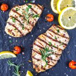 Two grilled chicken breasts garnished with rosemary and herbs on a dark surface, surrounded by grilled cherry tomatoes, coarse salt, and lemon slices.