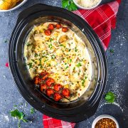 A creamy chicken casserole topped with roasted cherry tomatoes and chopped parsley is shown inside a black slow cooker, set on a gray surface with a red checkered napkin and dishes of seasonings nearby.