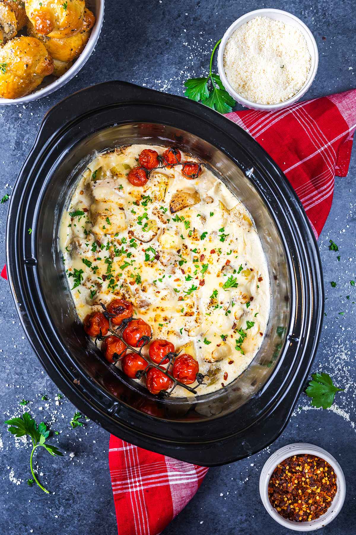 A slow cooker filled with creamy baked chicken, melted cheese, roasted cherry tomatoes, and chopped parsley, surrounded by bowls of potatoes, breadcrumbs, and red pepper flakes on a dark surface with a red napkin.